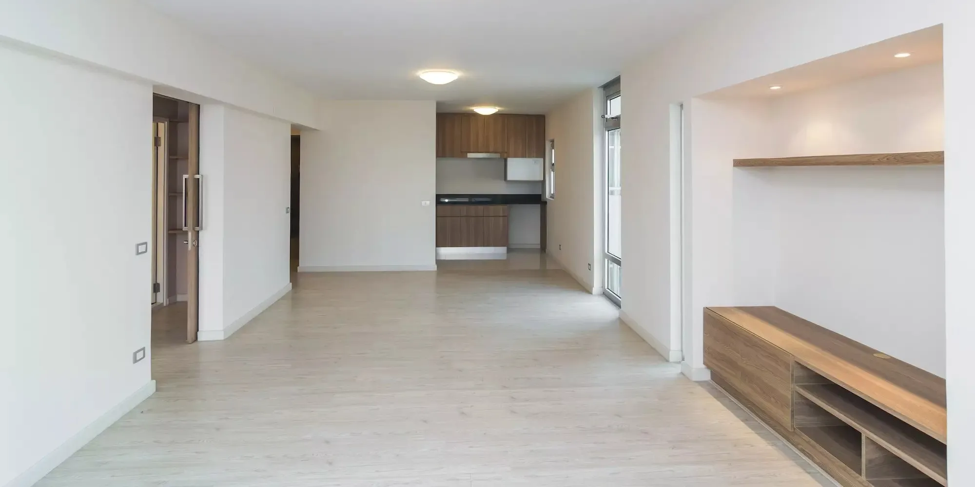 Empty bright condo unit with light wood floors, white walls, built-in shelving and an open kitchen